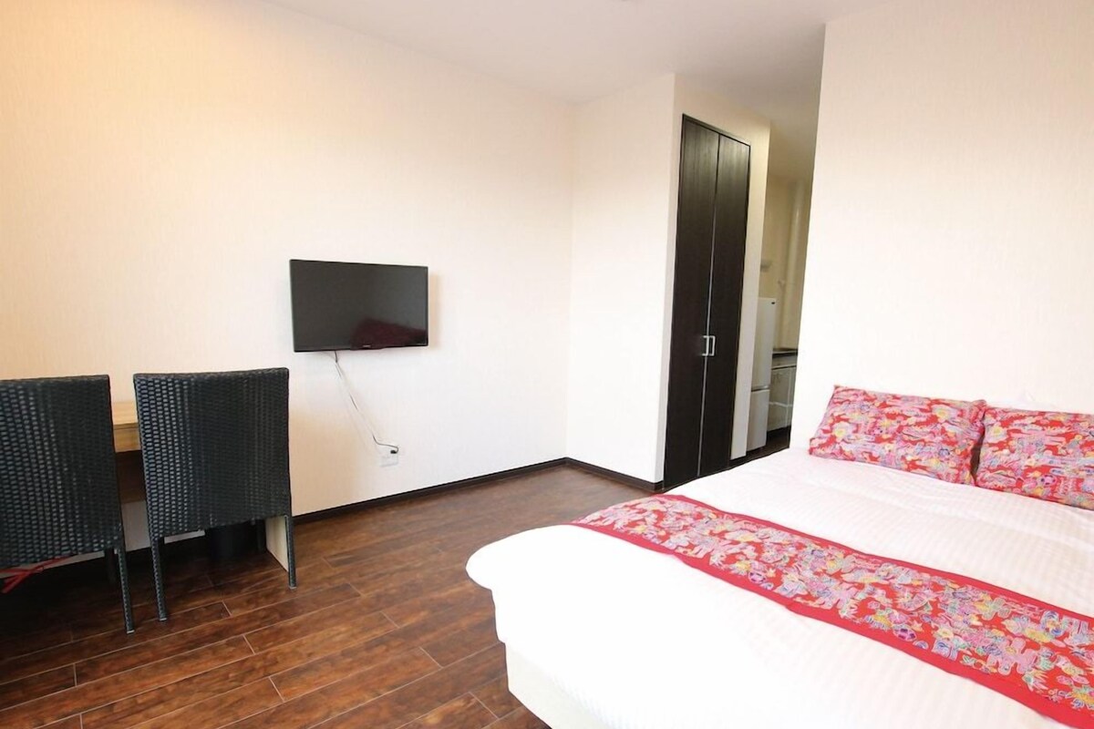 A hotel room with a bed, a TV mounted on the wall, a desk with a chair, and a wardrobe.