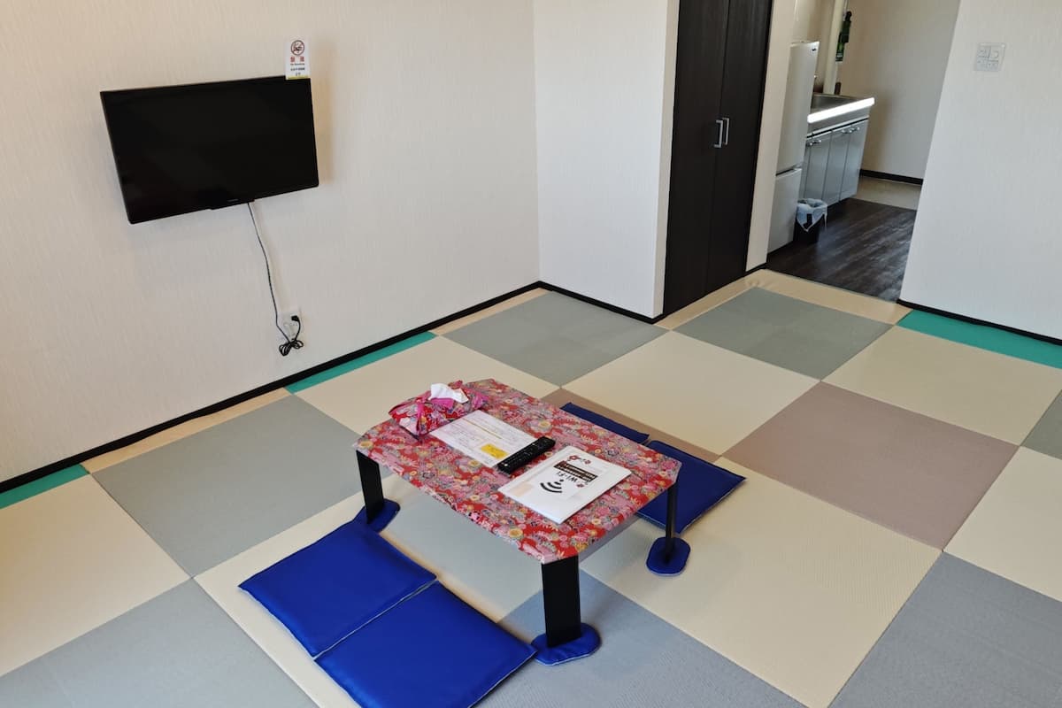 A small room with a flat-screen TV mounted on the wall, a small table with a floral tablecloth, and a blue cushioned bench.