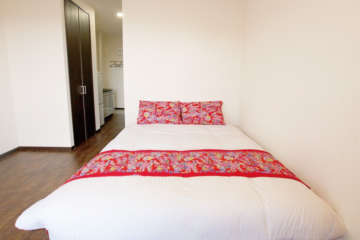 A bedroom with a bed, a wardrobe, and a small kitchenette in the background.