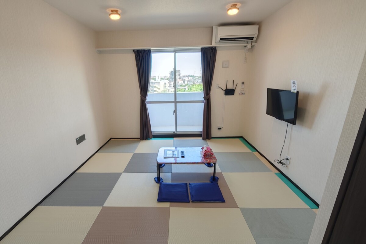 A hotel room with a checkered floor, a small table with a mat, a TV, and an air conditioning unit.