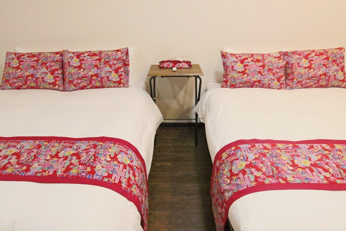 A hotel room with two beds, each with floral-patterned bedding and pillows. There is a small table with a red cloth and a white object on it.