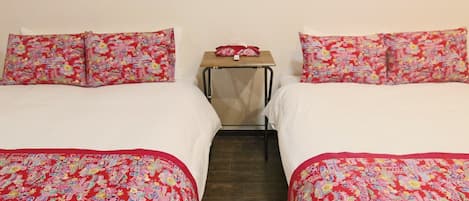 Una habitación de hotel con dos camas, cada una con ropa de cama y almohadas estampadas con motivos florales. Hay una mesita pequeña con un mantel rojo y un objeto blanco encima.