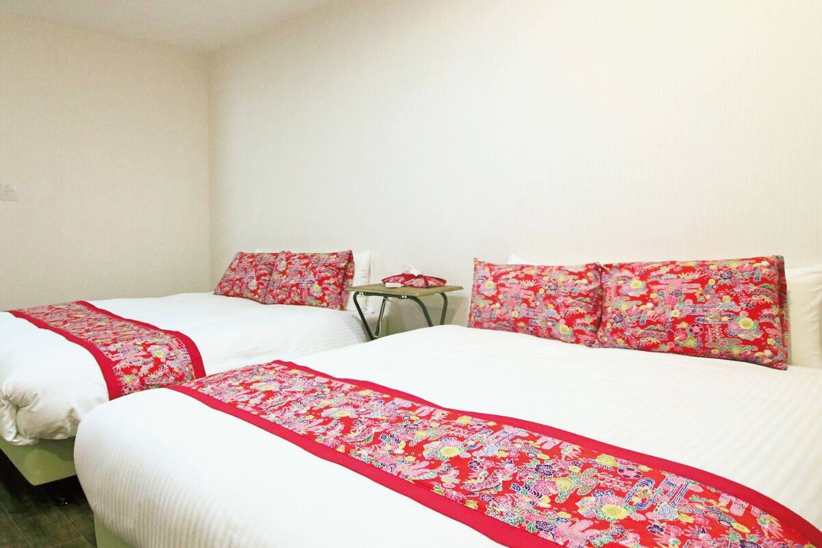 A hotel room with two beds, each with white bedding and red floral bedspreads. There is a small table with a chair in the room.