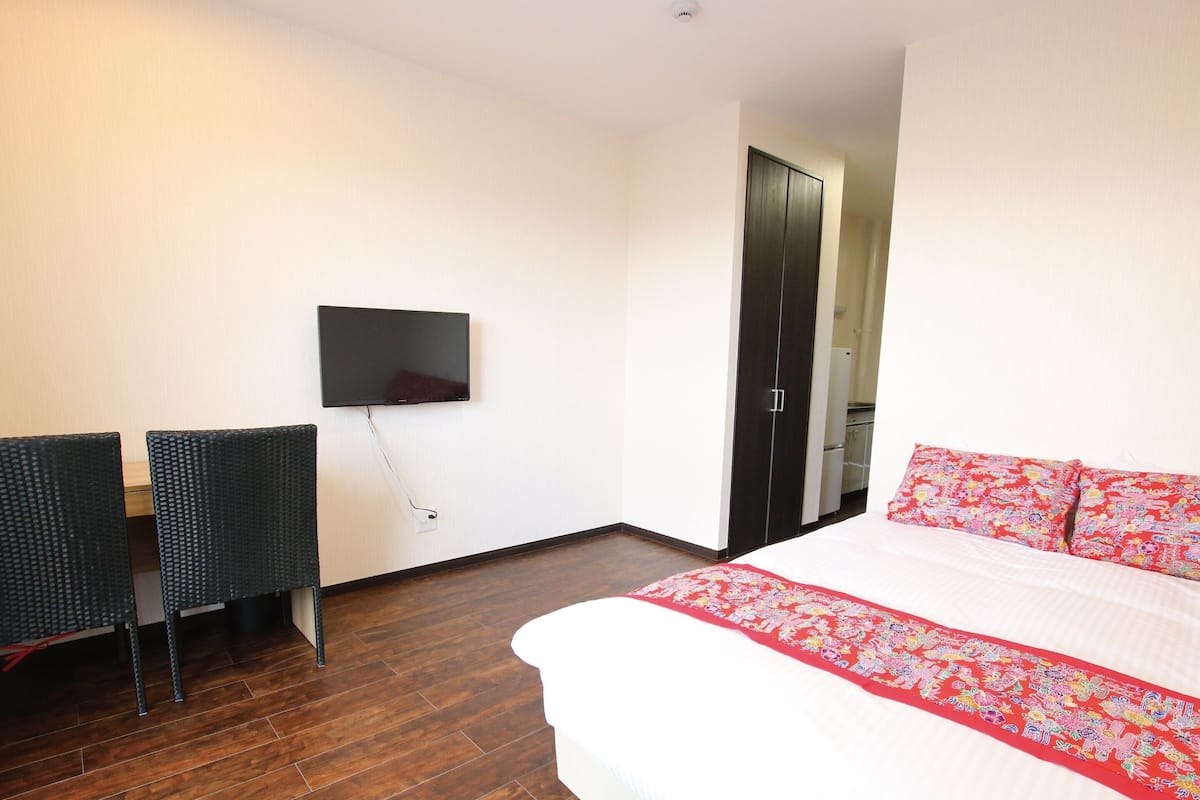 A hotel room with a bed, a TV mounted on the wall, a desk with two chairs, and a wardrobe.