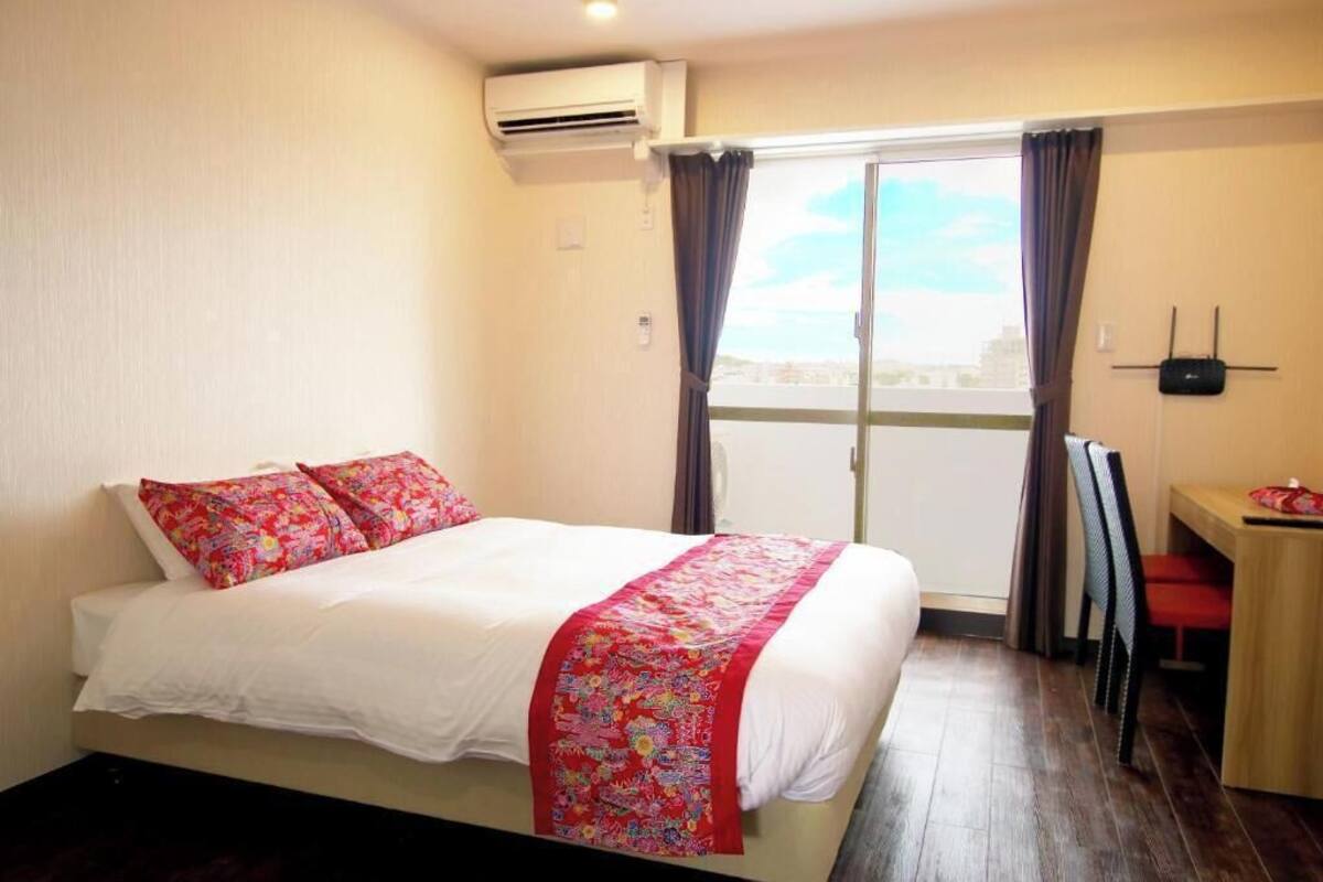 A hotel room with a bed, a desk, a chair, an air conditioning unit, a window with curtains, and a view of the city.