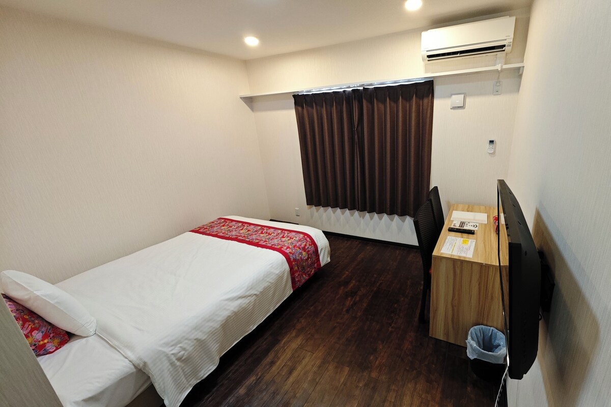 A small, clean room with two single beds, a wooden desk, a chair, an air conditioning unit, and a small trash bin.