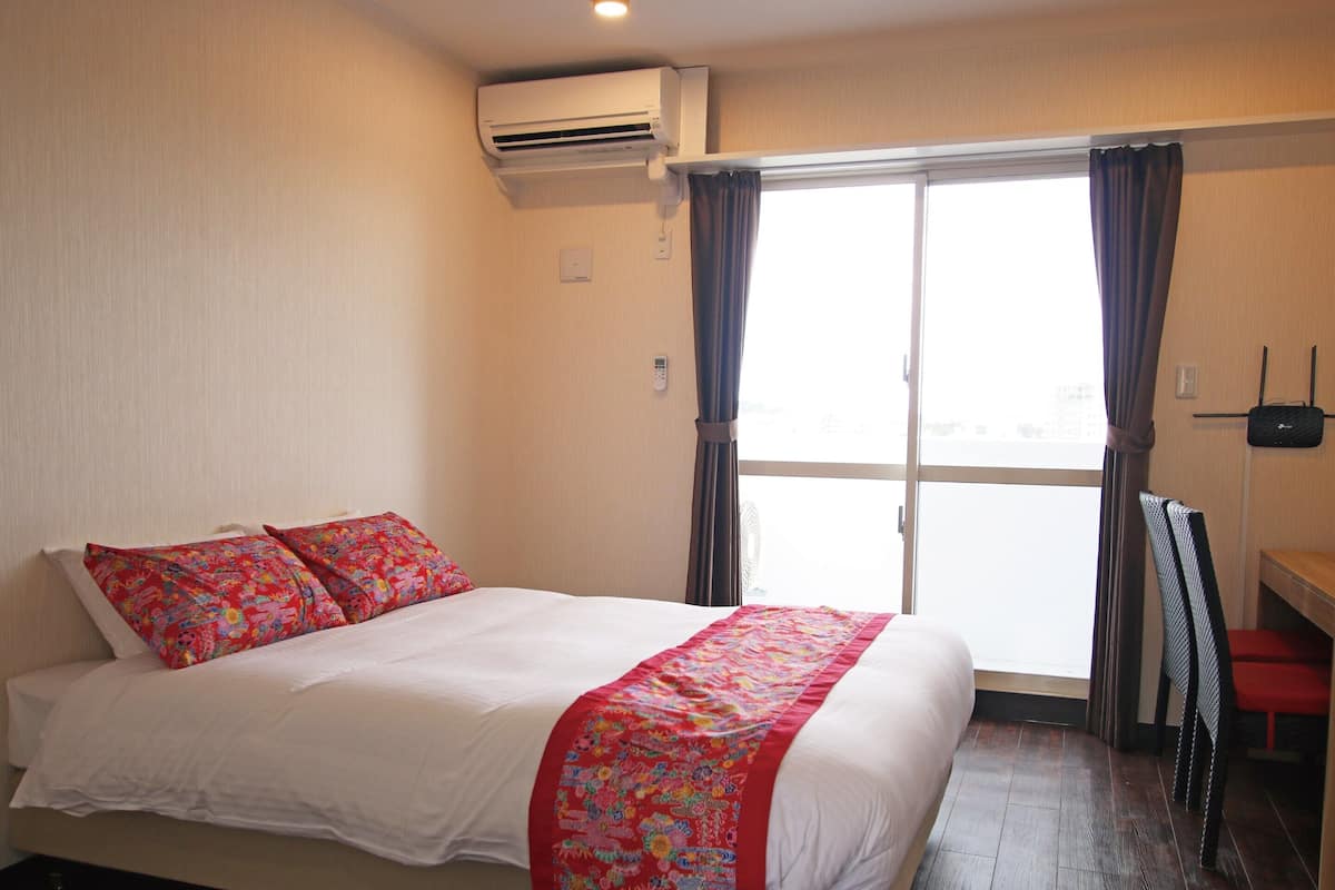 A hotel room with a bed, a window with curtains, a ceiling fan, and a wall-mounted device.