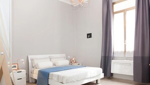 A modern bedroom with a white bed, a nightstand, a bedside lamp, a window with curtains, and a radiator. - il Nespolo (Bologna)