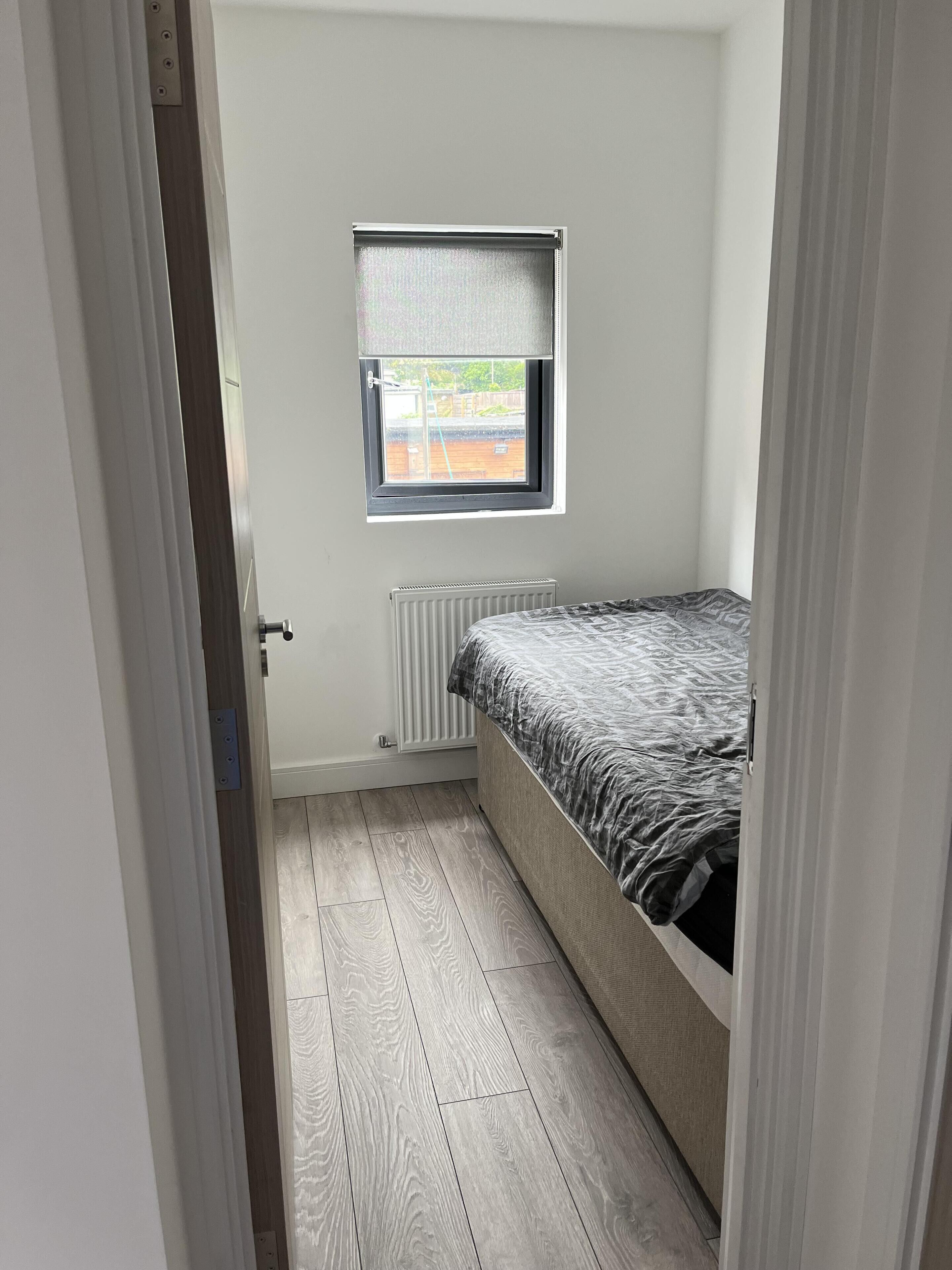 A small, narrow room with a single bed, a window with a blind, and a radiator on the wall.