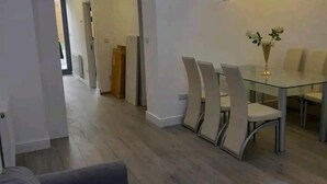A modern dining area with a glass table and white chairs, a grey sofa, and a hallway leading to another room. - Apartment - Sleeps up to 6 & Free Parking (London)