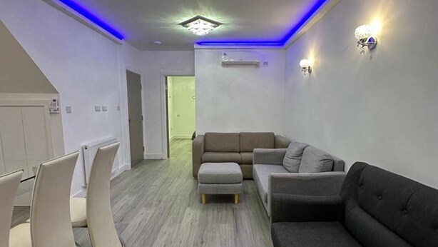 A modern living room with a grey sofa, a grey armchair, and a small ottoman. There is a dining area with white chairs and a table. The room has a wooden floor and a ceiling with blue LED lighting. - Apartment - Sleeps up to 6 & Free Parking (London)