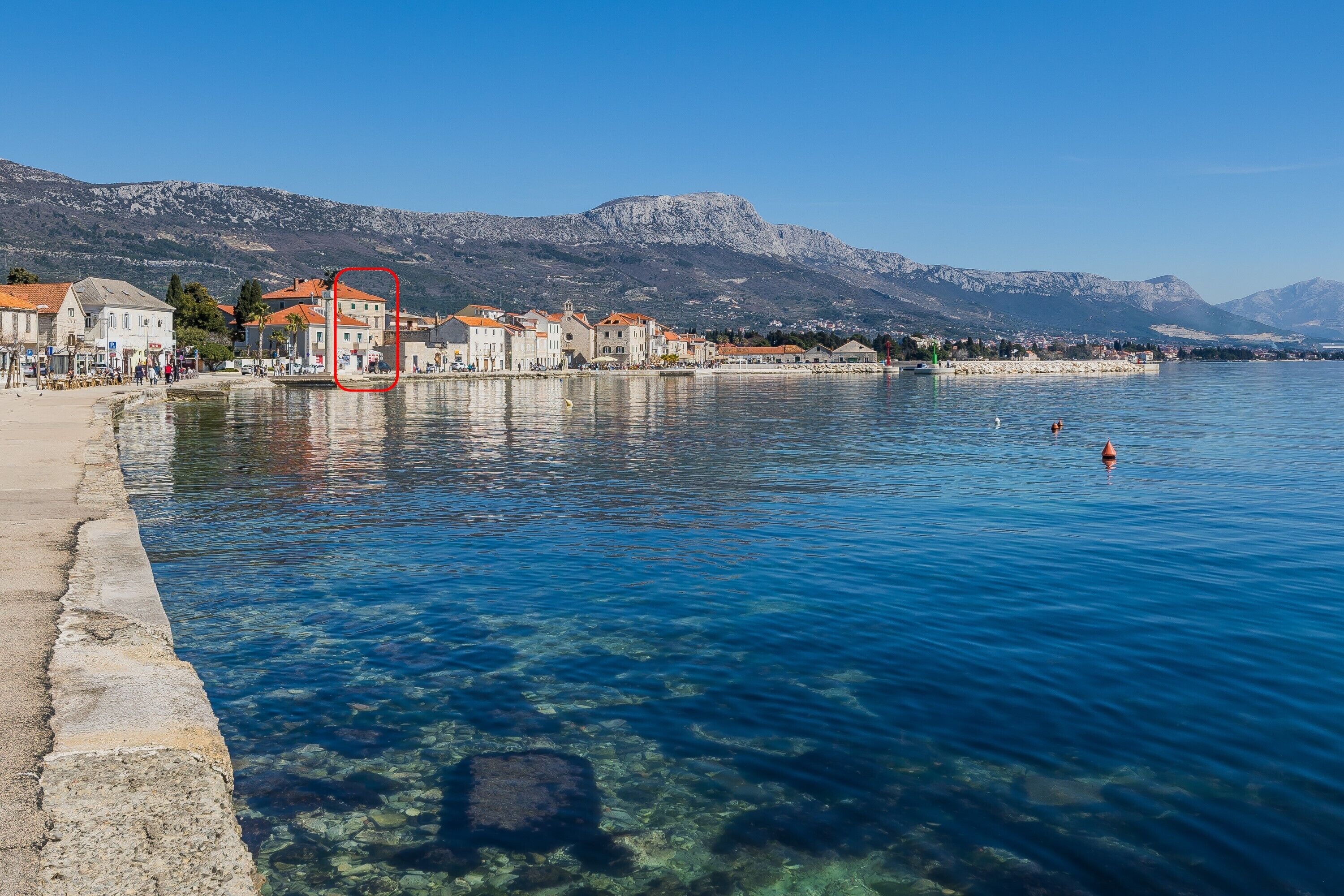 Un hôtel situé au bord de l’eau, avec une eau d’un bleu limpide, une jetée en pierre et des montagnes en arrière-plan.