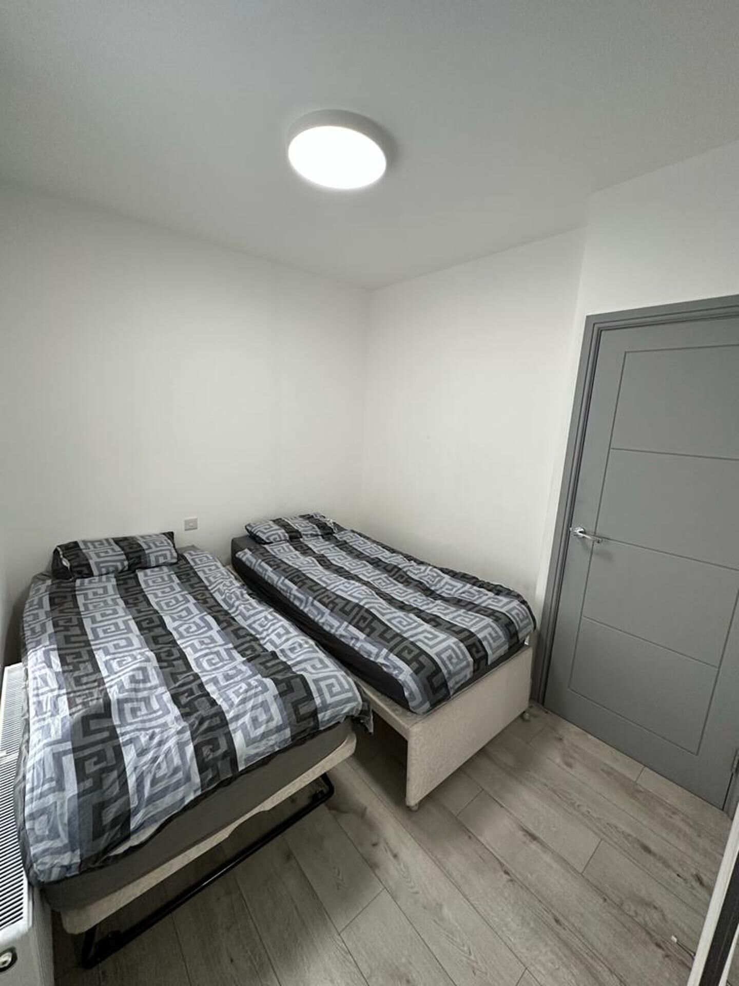 A small, single-room accommodation with two single beds, a built-in bench, a door, and a ceiling light.