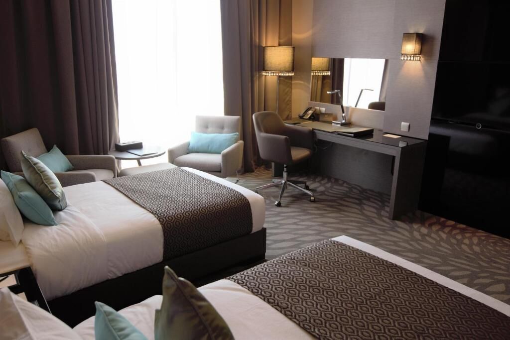 Quarto de hotel com uma cama grande, duas poltronas, uma escrivaninha com computador, um espelho e uma televisão.