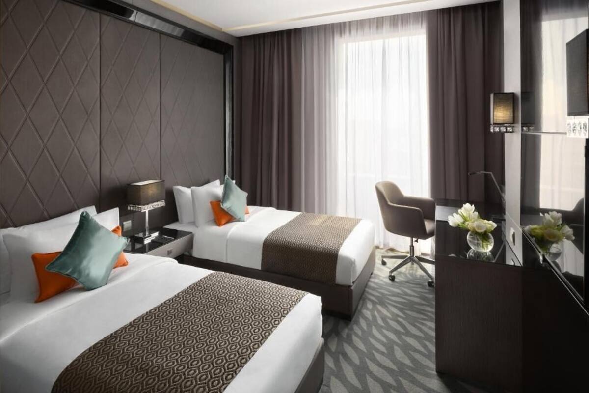 Habitación de hotel con dos camas, un escritorio, una silla y un amplio ventanal con cortinas.