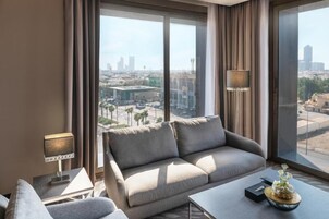 Habitación de hotel moderna con un ventanal grande que ofrece vistas a la ciudad, un sofá, una mesita y una lámpara.