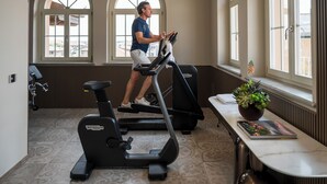 A man using an elliptical machine in a room with large arched windows, a radiator, and a table with a plant and a magazine. - Roof Suite - Castello il Codirosso Accomodation Hotel (Cesate)
