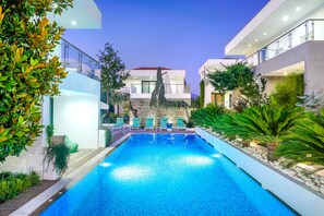 Un área de piscina moderna con una piscina azul cristalina, sillas de descanso verdes y un muro de piedra de fondo.