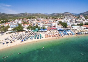 Un pueblo costero con una playa bordeada de sombrillas y tumbonas, un mar azul cristalino y edificios al fondo.