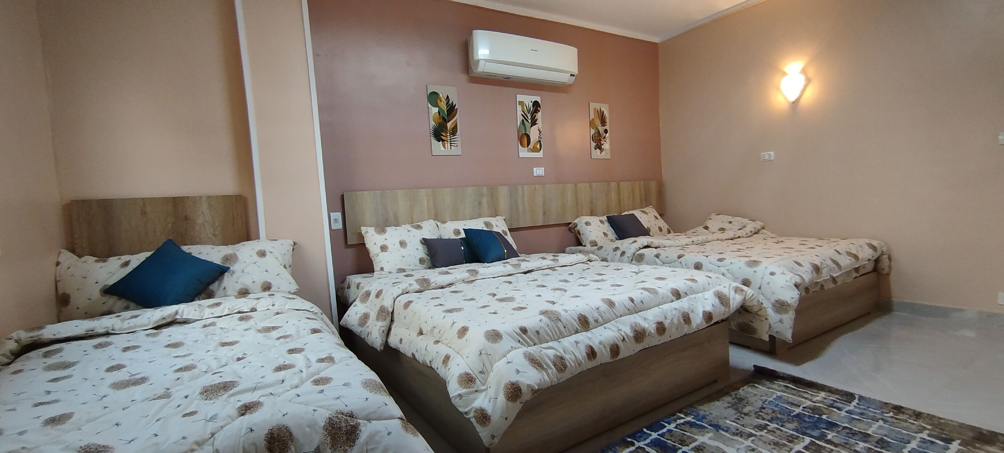 Habitación de hotel con tres camas, cada una con cabecero de madera y ropa de cama estampada. Se observa un aire acondicionado de pared y una obra de arte en la pared sobre las camas.