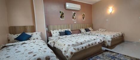 A hotel room with three beds, each with a wooden headboard and patterned bedding. There is a wall-mounted air conditioner and wall art above the beds.