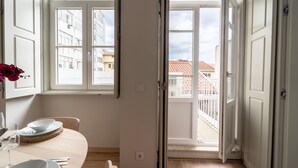 Un comedor con una mesa preparada para dos personas, una ventana con vistas a edificios y una puerta abierta que da al exterior.