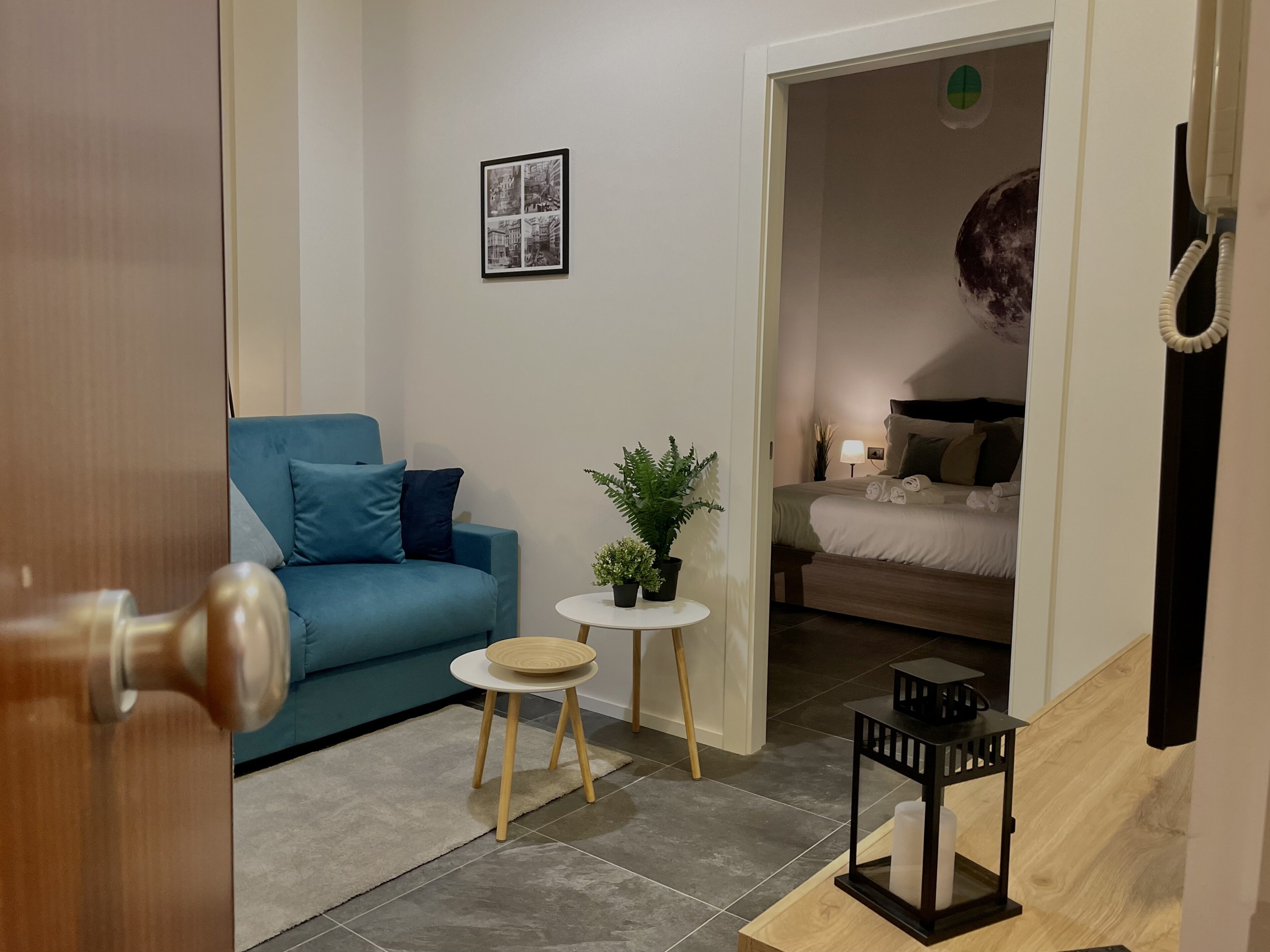 A modern living room with a blue sofa, a small round table with a plant, and a bed in the background.