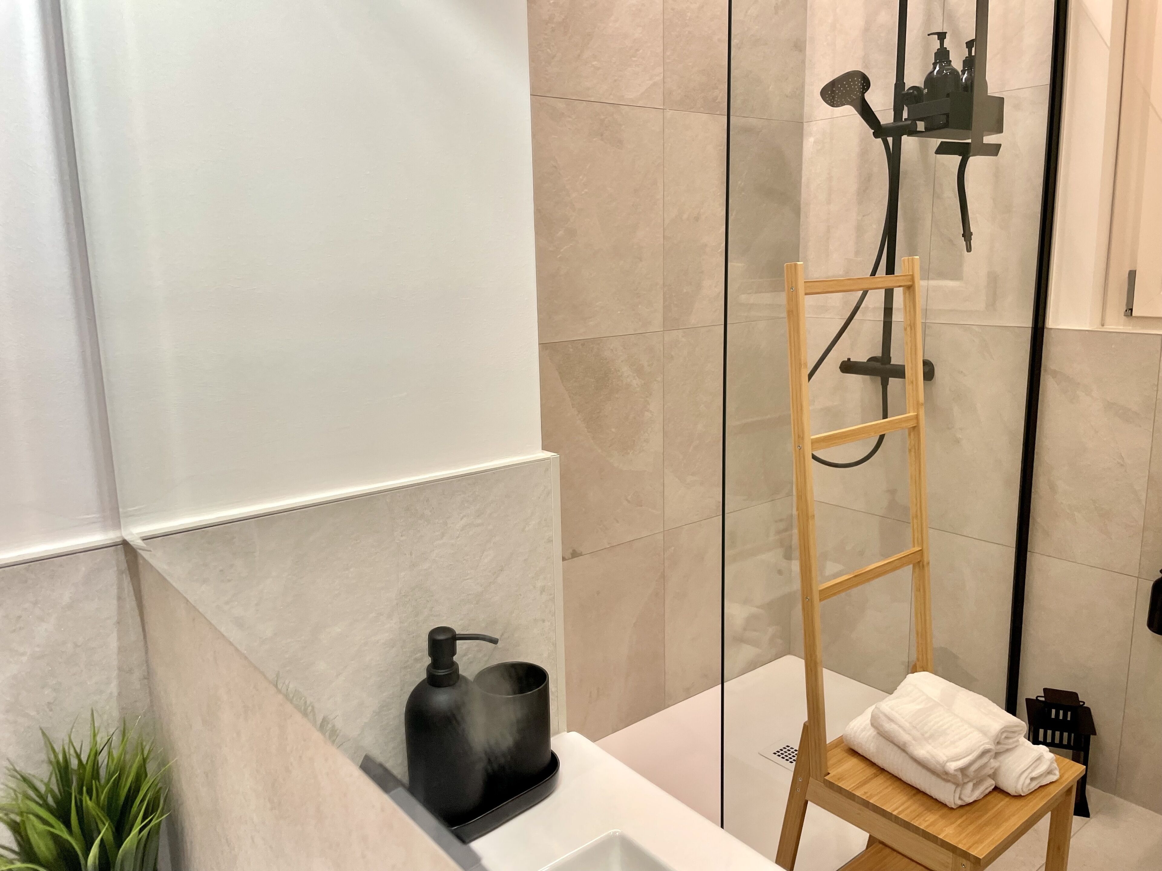 A modern bathroom with a glass shower enclosure, a wooden ladder, a white sink, and a small wooden table with towels.