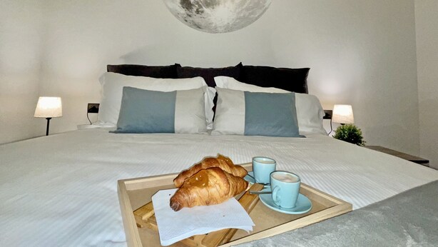 Quarto de hotel com cama, almofadas, bandeja com croissants e xícaras de café, e um mural na parede retratando a lua.