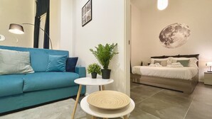 Quarto moderno e bem iluminado, com um sofá azul, uma mesa redonda com uma planta, uma cama com roupa de cama bem arrumada e uma luminária fixada na parede.