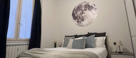 A bedroom with a bed, a nightstand, a lamp, a window with curtains, and a wall mural of the moon.