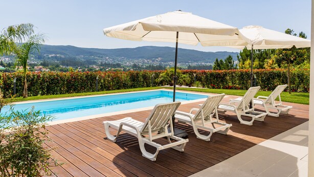 Zona de piscina con sillones blancos y sombrillas, rodeada de vegetación y vistas a colinas distantes.