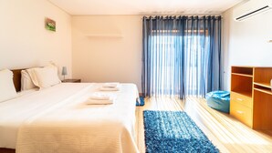 A hotel room with a large bed, a blue rug, a wooden wardrobe, and a window with sheer curtains.
