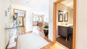 A modern interior with a wooden floor, a white rug, a glass console table, a dark-toned bathroom with a mirror, and a potted plant.