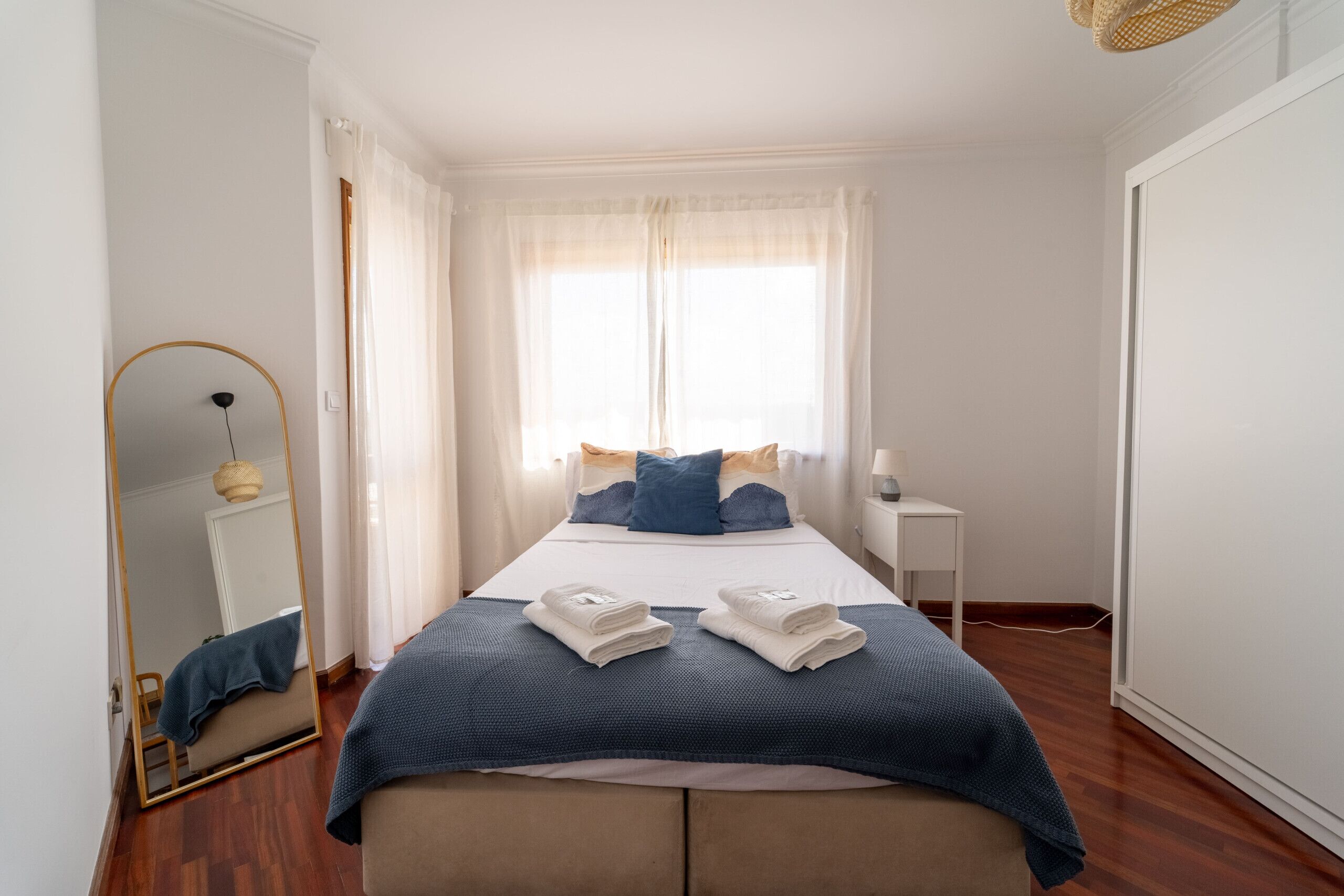 A neatly arranged hotel room with a bed, a bedside table, a mirror, and a window with curtains.