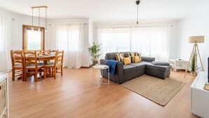 Ein modernes Wohnzimmer mit einem grauen Ecksofa, einem Holzesstisch mit Stühlen, einem kleinen Beistelltisch und einem großen Fenster mit durchsichtigen Vorhängen.