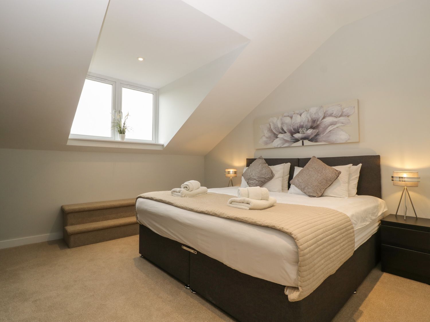 A bedroom with a large bed, a skylight, a painting, and a nightstand with a lamp.