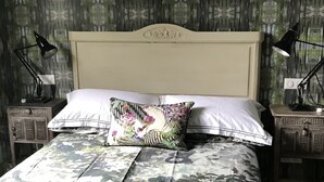 Una cama con una colcha de estampado floral y una almohada decorativa, flanqueada por dos mesitas de noche con lámparas, frente a una pared cubierta de papel tapiz.