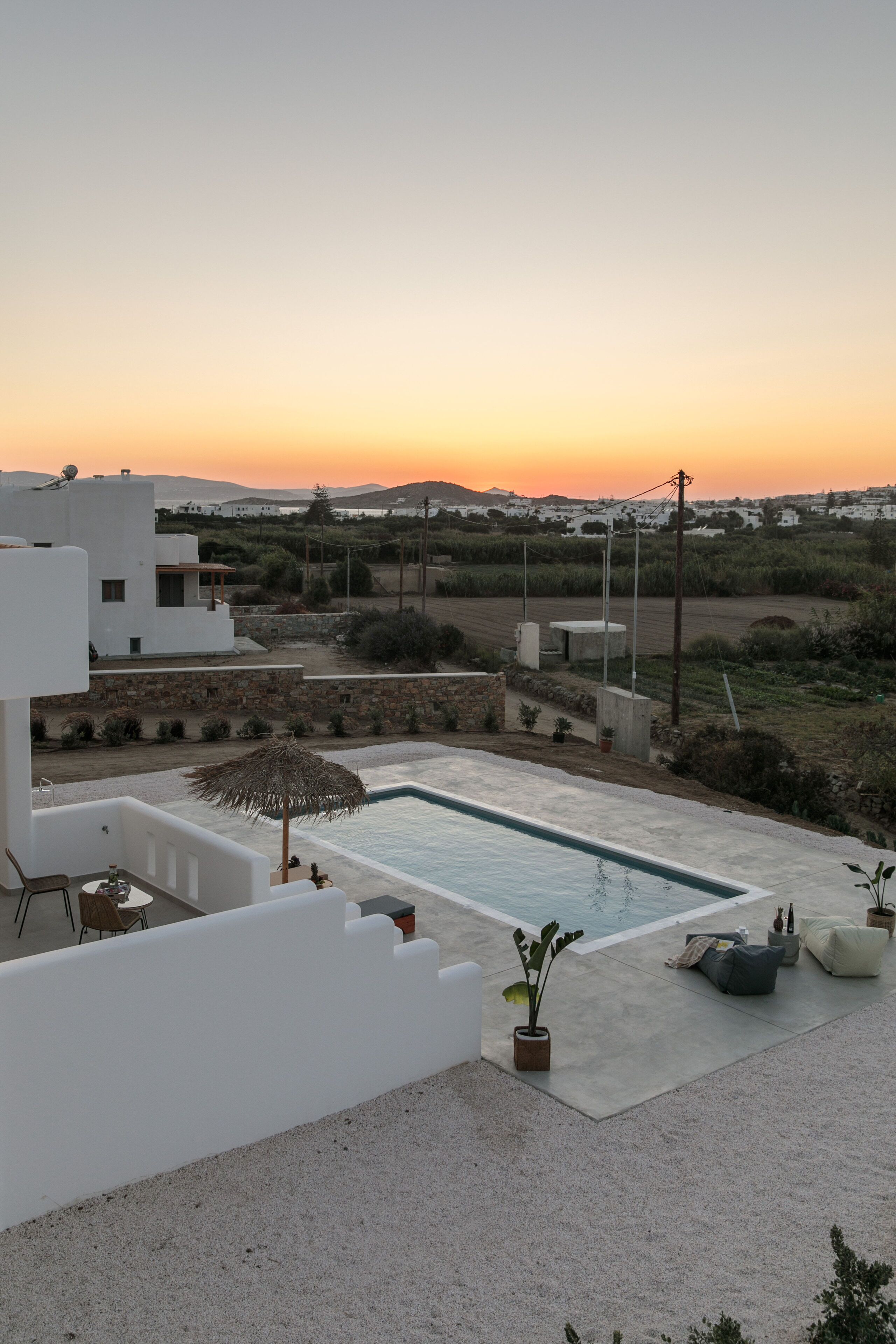 A modern outdoor pool area with a clear view of the sunset, surrounded by white buildings and a grassy landscape.