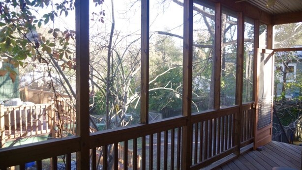 Una terraza de madera con barandilla, ventanas amplias y vistas a árboles y una valla.