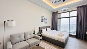 Une chambre d’hôtel moderne, dotée d’un grand lit, d’un canapé, d’une lampe sur pied et offrant une vue sur la ville grâce à de grandes fenêtres.