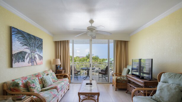 A living room with a sofa, coffee table, and wicker chairs, a television, and a balcony with a view of palm trees. - Sunrise 307-2BR/2BA condo near beaches with pool, parking, balcony (Key West)