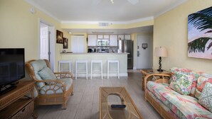 A living room with a television, wicker furniture, a sofa, and a kitchen area with a bar. - Sunrise 307-2BR/2BA condo near beaches with pool, parking, balcony (Key West)