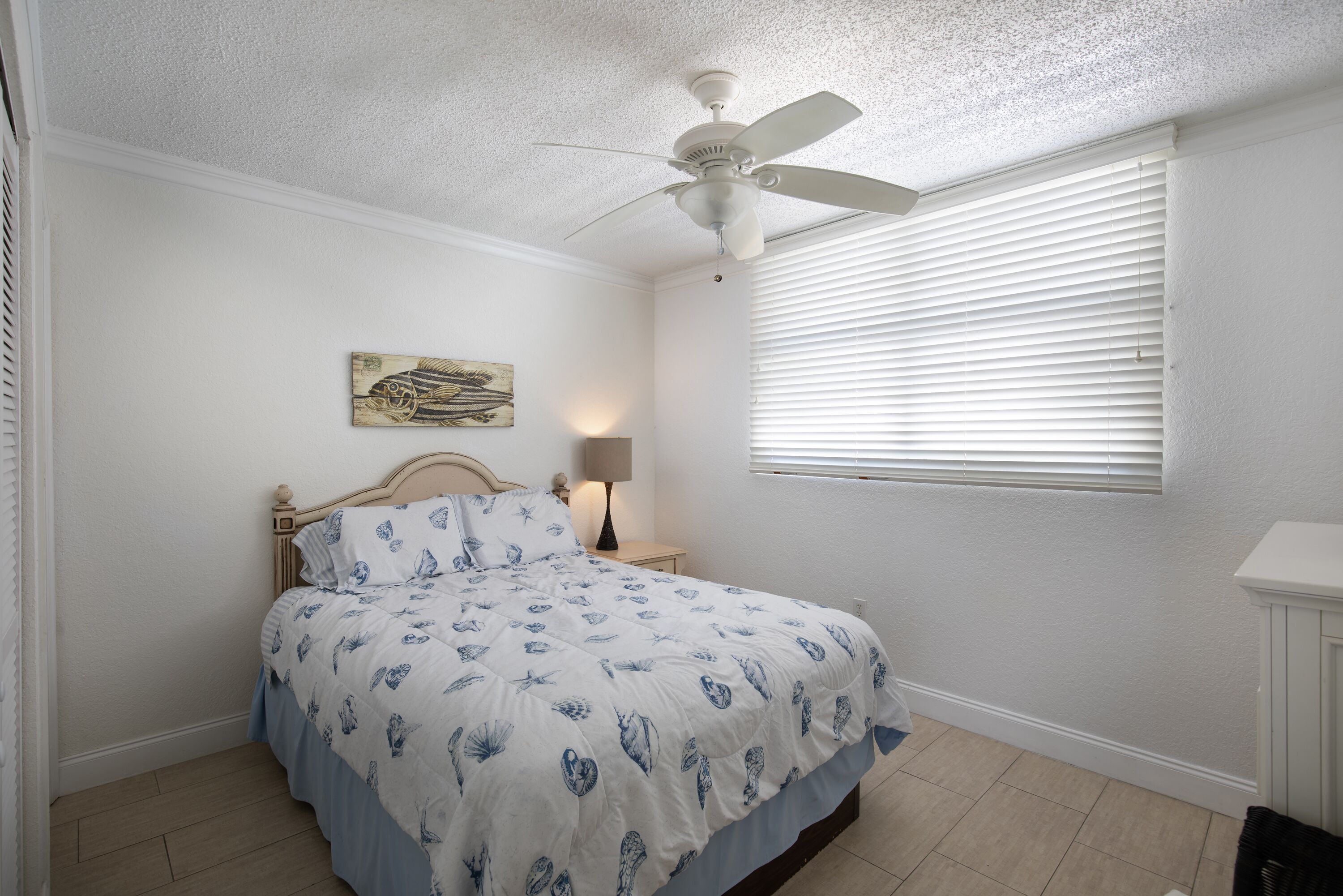 A bedroom with a bed, a ceiling fan, a window with blinds, a bedside lamp, and a wall-mounted artwork.