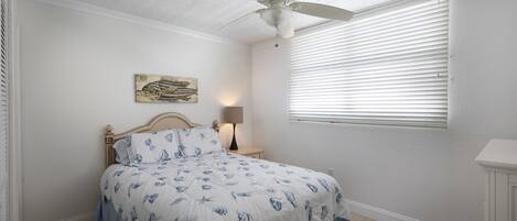 A bedroom with a bed, a ceiling fan, a window with blinds, a bedside lamp, and a wall-mounted artwork.