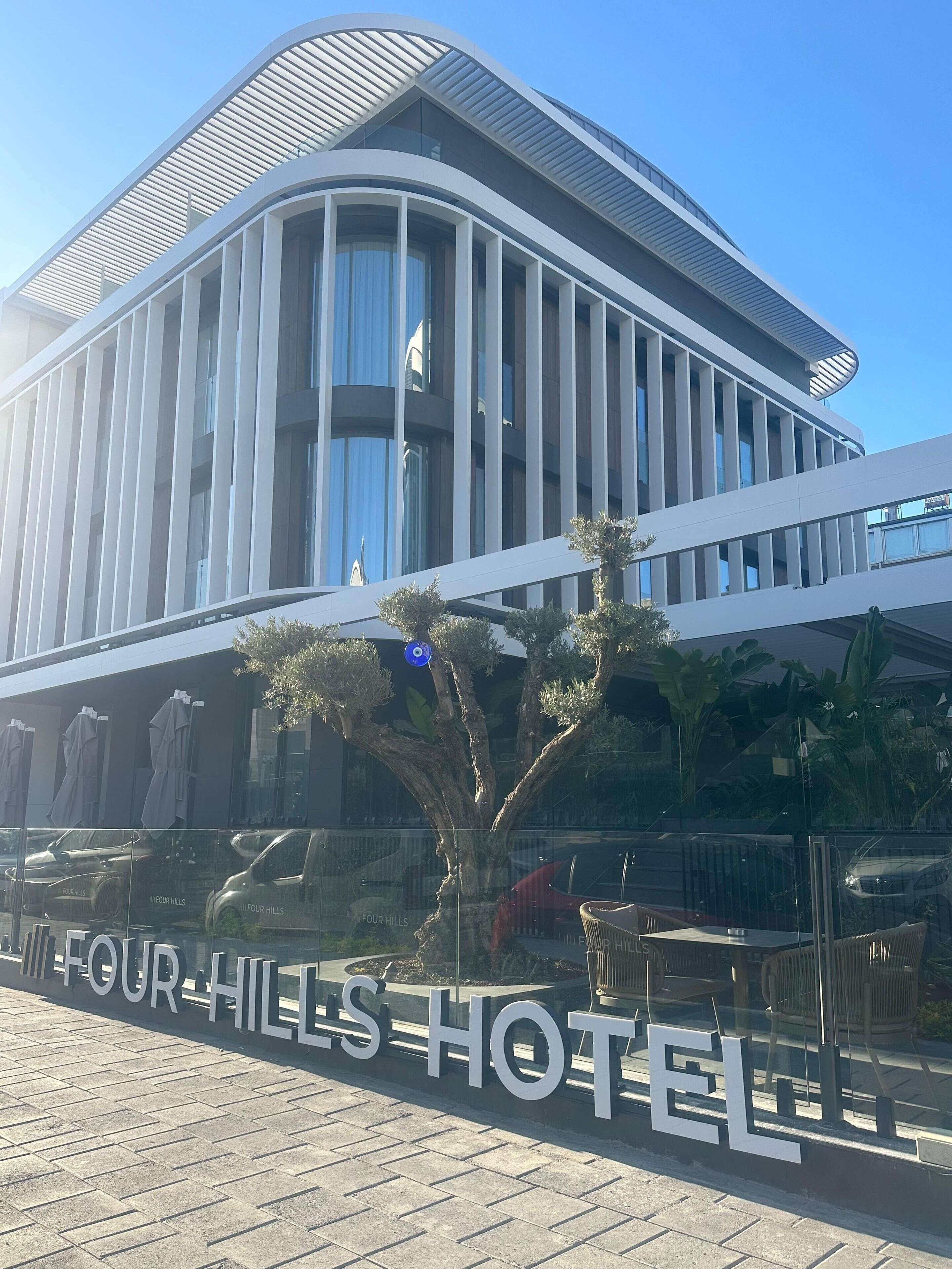 Een modern hotel met een gebogen gevel, grote ramen en een bord waarop 'FOUR HILLS HOTEL' staat.