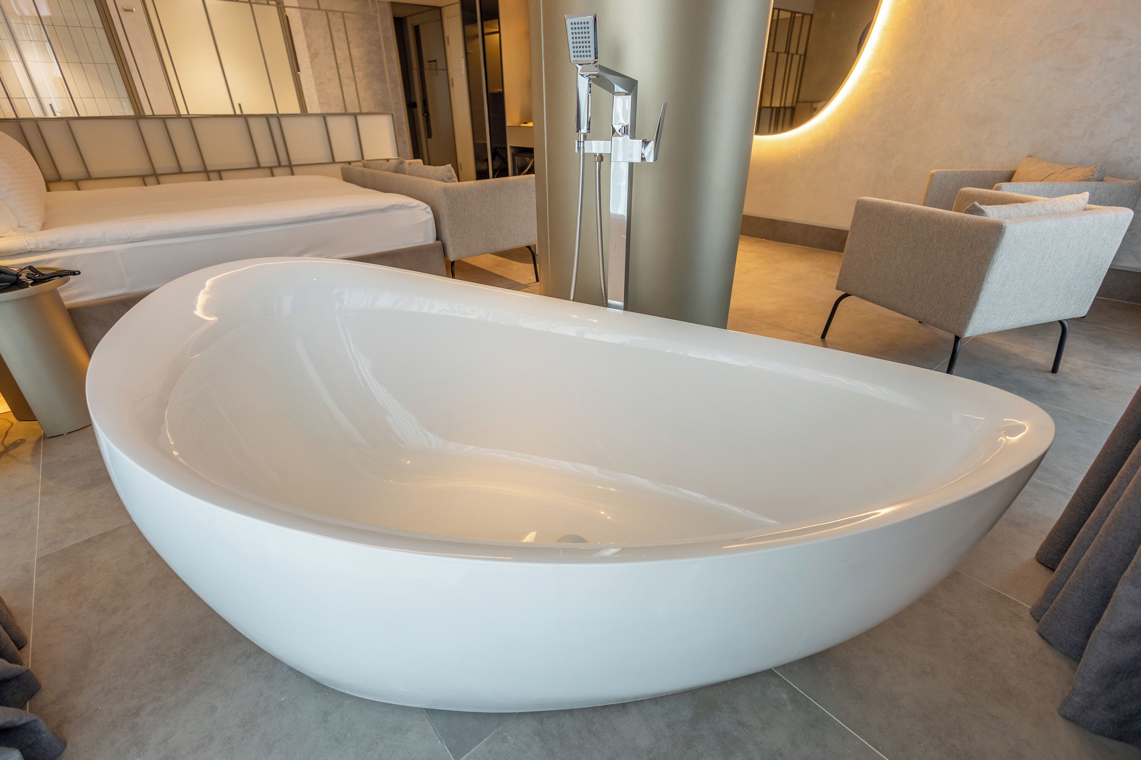 A modern bathroom with a large oval bathtub, a seating area with a sofa and armchairs, and a wall-mounted showerhead.