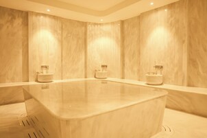 Una bañera de mármol rectangular grande con dos inodoros a cada lado, ubicada en una habitación con paredes de mármol alto y luces empotradas.