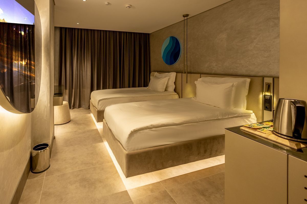 A modern hotel room with two beds, a large window with curtains, a wall-mounted artwork, and a small kitchenette area.