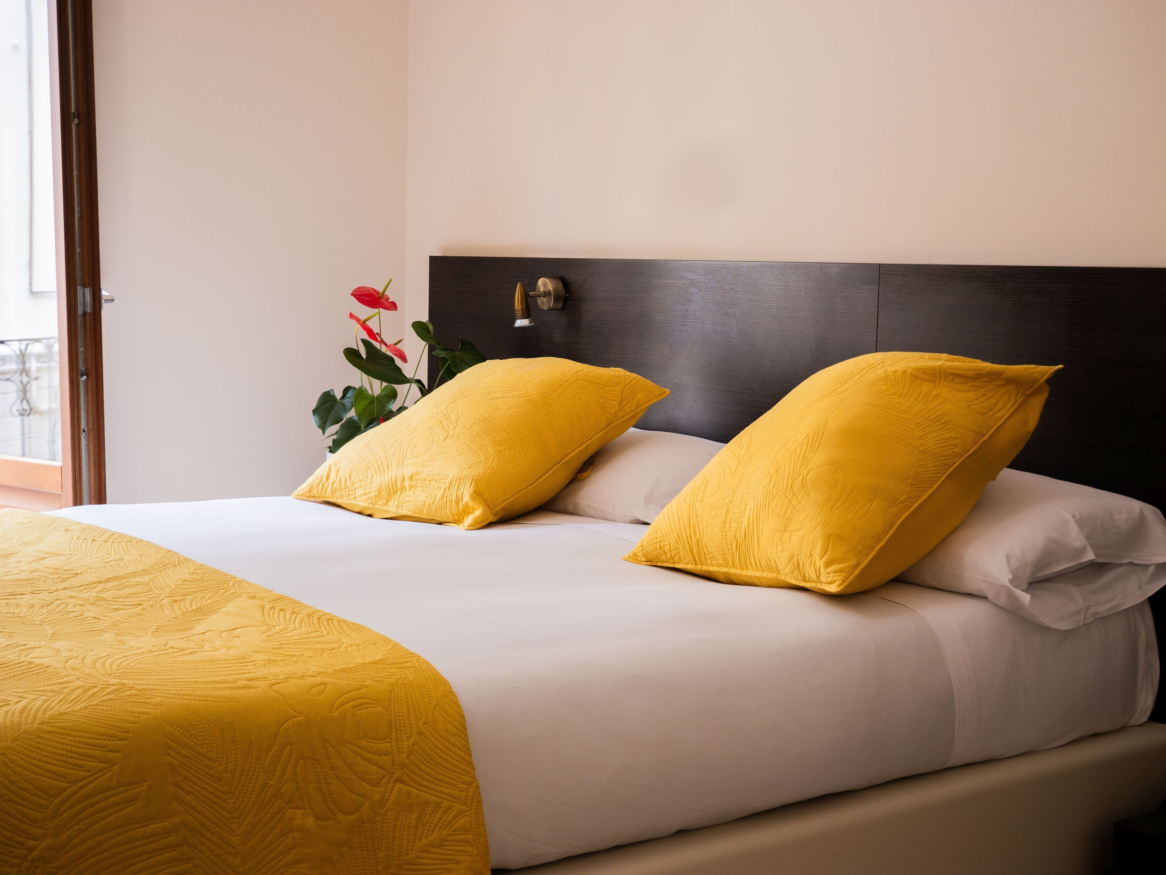 A hotel room with a large bed, two yellow pillows, a wooden headboard, and a potted plant with red flowers.